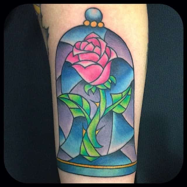 A colorful Beauty and the Beast-inspired tattoo of a pink rose inside a stained glass dome with vibrant blue and green accents
