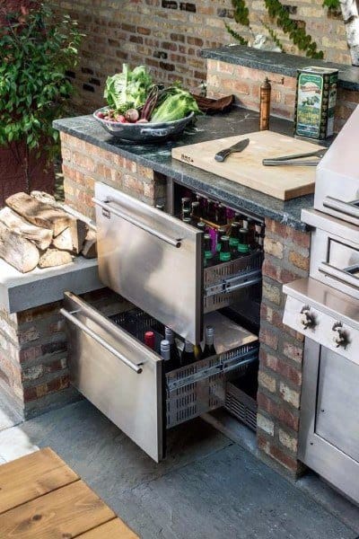 Outdoor kitchen featuring a brick countertop, open stainless drawers with beverages, and fresh vegetables on top