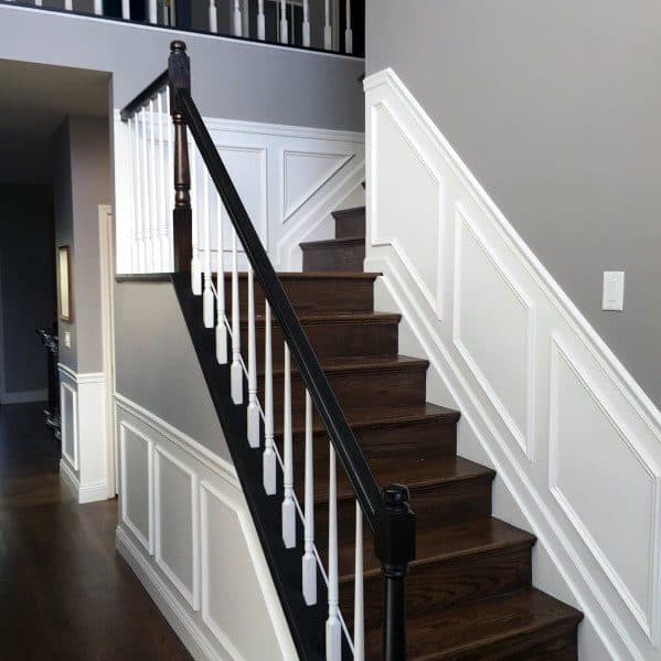 A wooden staircase with dark steps, white balusters, and chair rail accenting the gray wainscoting panels
