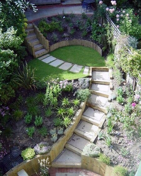 Beautiful sloped garden with wooden stairs, a curved pathway, lush greenery, and a small fenced lawn area for a cozy outdoor space