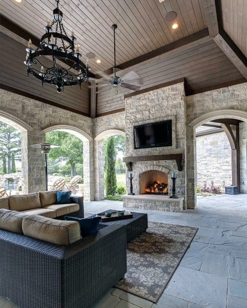 A cozy patio features a stone fireplace, TV, chandelier, and cushioned wicker furniture surrounded by lush greenery