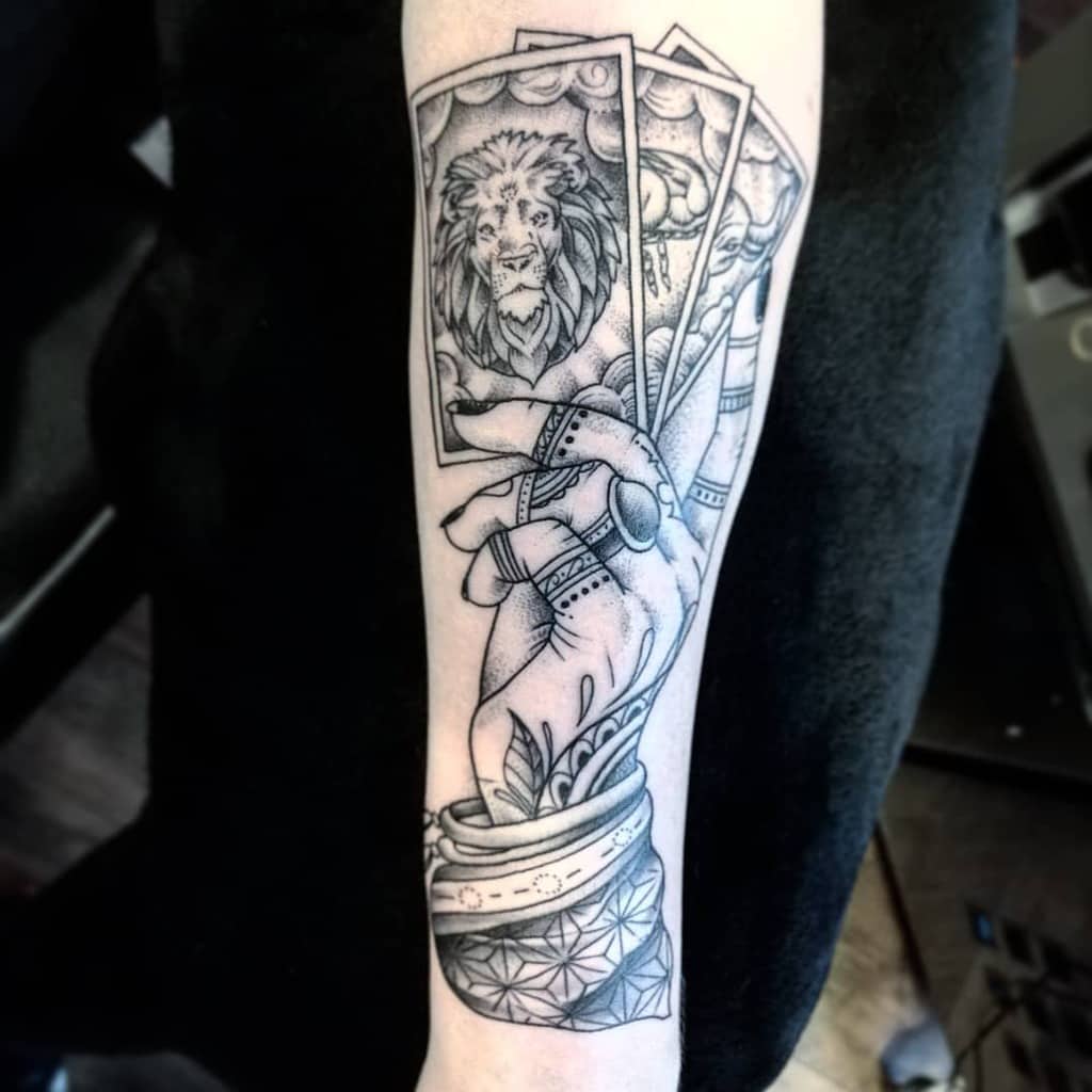 Black and grey tattoo of a mystical hand holding tarot cards, featuring a lion, celestial elements, and intricate shading on the forearm.