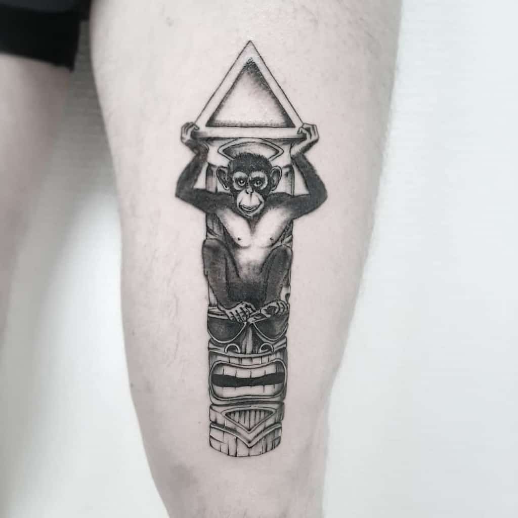 Black and grey tattoo featuring a Tiki mask with a monkey sitting on top, holding a triangular shape