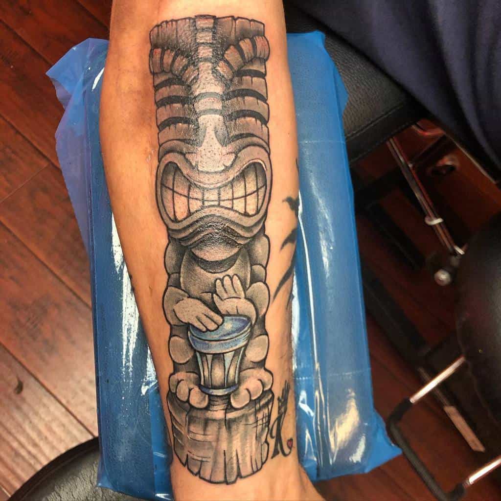 Black and grey Tiki tattoo on the forearm, featuring a grinning Tiki mask character drumming on a drum