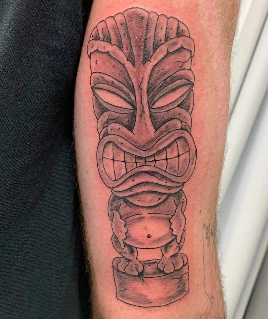 Black and grey Tiki tattoo featuring a detailed Tiki mask with bold shading, standing on a pedestal