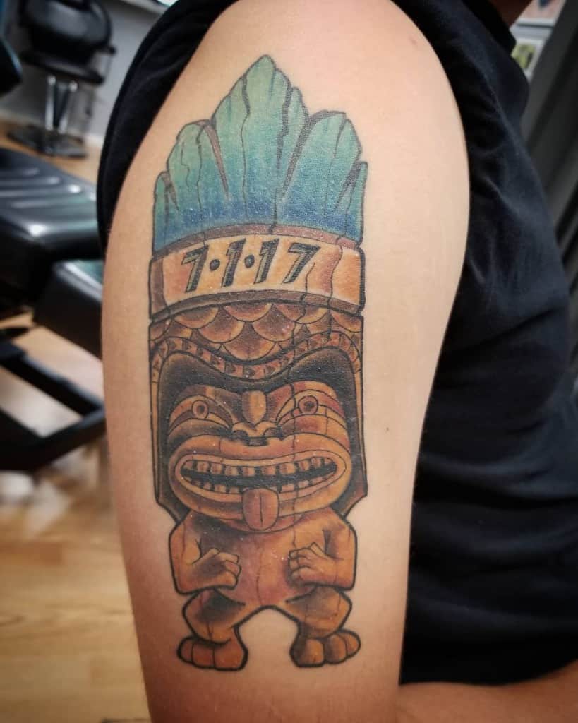 Colorful Tiki tattoo featuring a smiling Tiki mask with a blue and green feathered crown, and the date '7:1:17' inscribed on the top