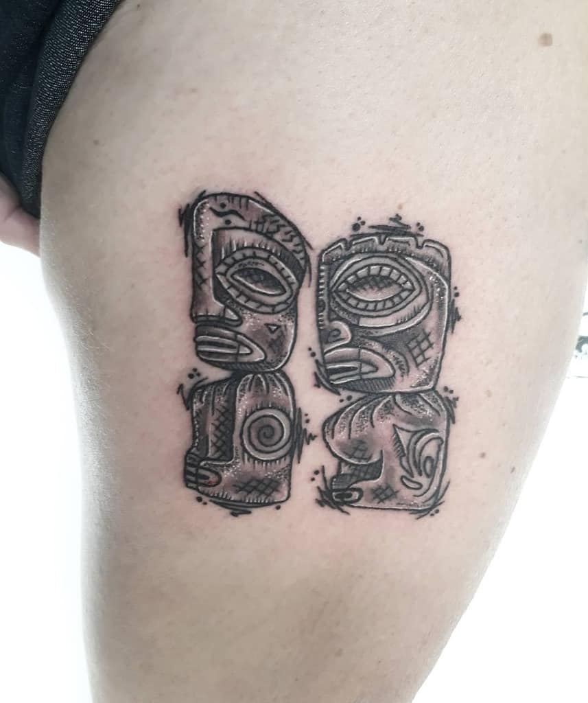 Black and grey Tiki tattoo featuring two stylized Tiki masks with intricate lines, geometric patterns, and subtle shading, positioned next to each other