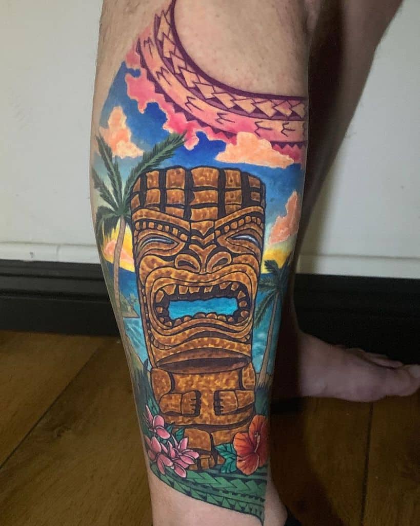 Vibrant Tiki tattoo featuring a Tiki mask with a colorful sunset background, ocean, palm trees, and tropical flowers with green leaves