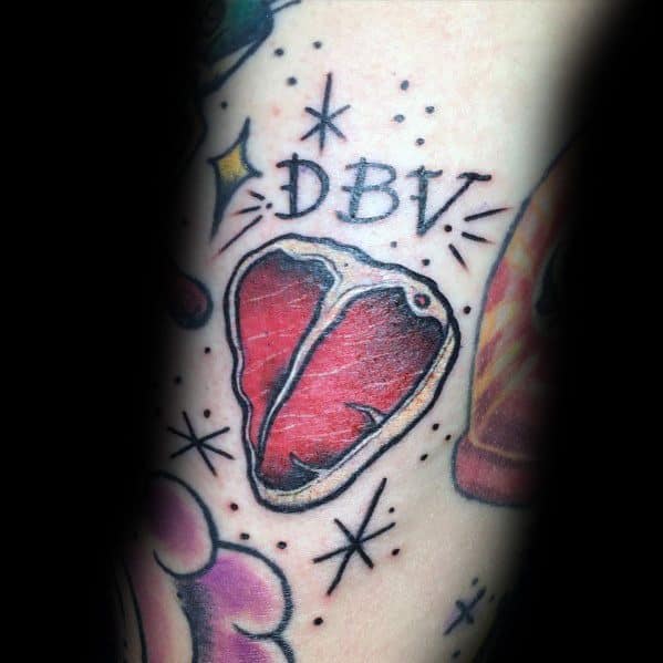 Tattoo of a heart-shaped steak with bold black outlines, red shading, and the letters DBV above, surrounded by sparkles for a classic design