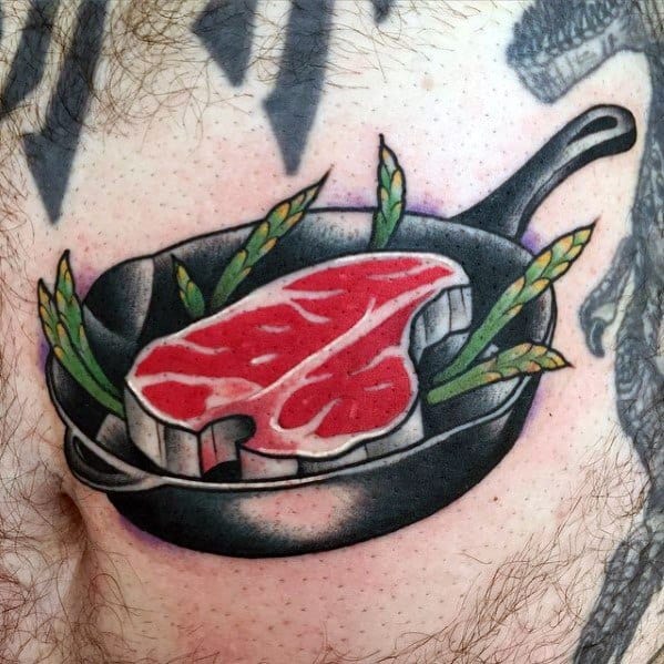 Tattoo of a raw steak in a cast-iron pan with asparagus, featuring bold shading and vibrant colors for a detailed cooking-inspired design
