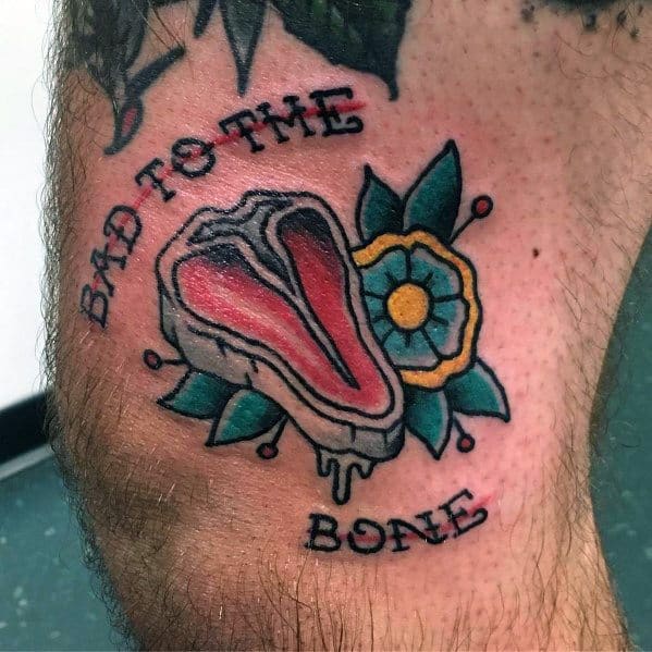 Tattoo of a steak with dripping details, surrounded by a flower, featuring the phrase "Bad to the Bone" in bold lettering for a classic design.