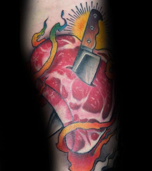 Tattoo of a raw steak shaped like a heart, pierced by a cleaver, with flames and a radiant sunburst for a bold and intense design.