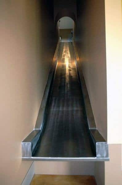 A sleek, metallic indoor slide gracefully descends between two walls, with a small arched entrance visible at the top