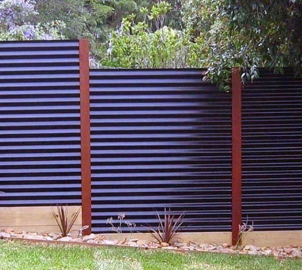 Corrugated metal fence with wooden posts, surrounded by lush plants and stones in a serene garden setting