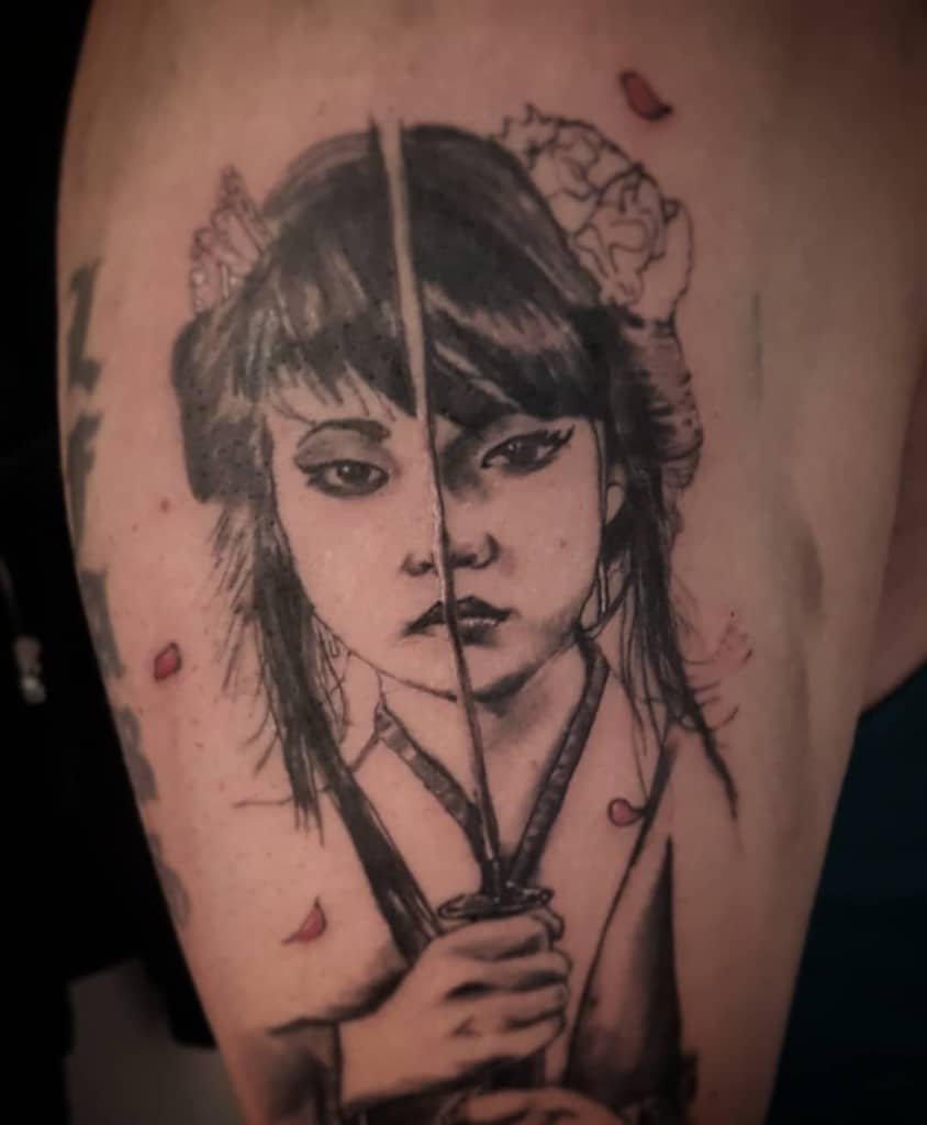 Black and grey tattoo of a fierce Geisha holding a katana, with the blade splitting her face symmetrically, surrounded by falling petals.
