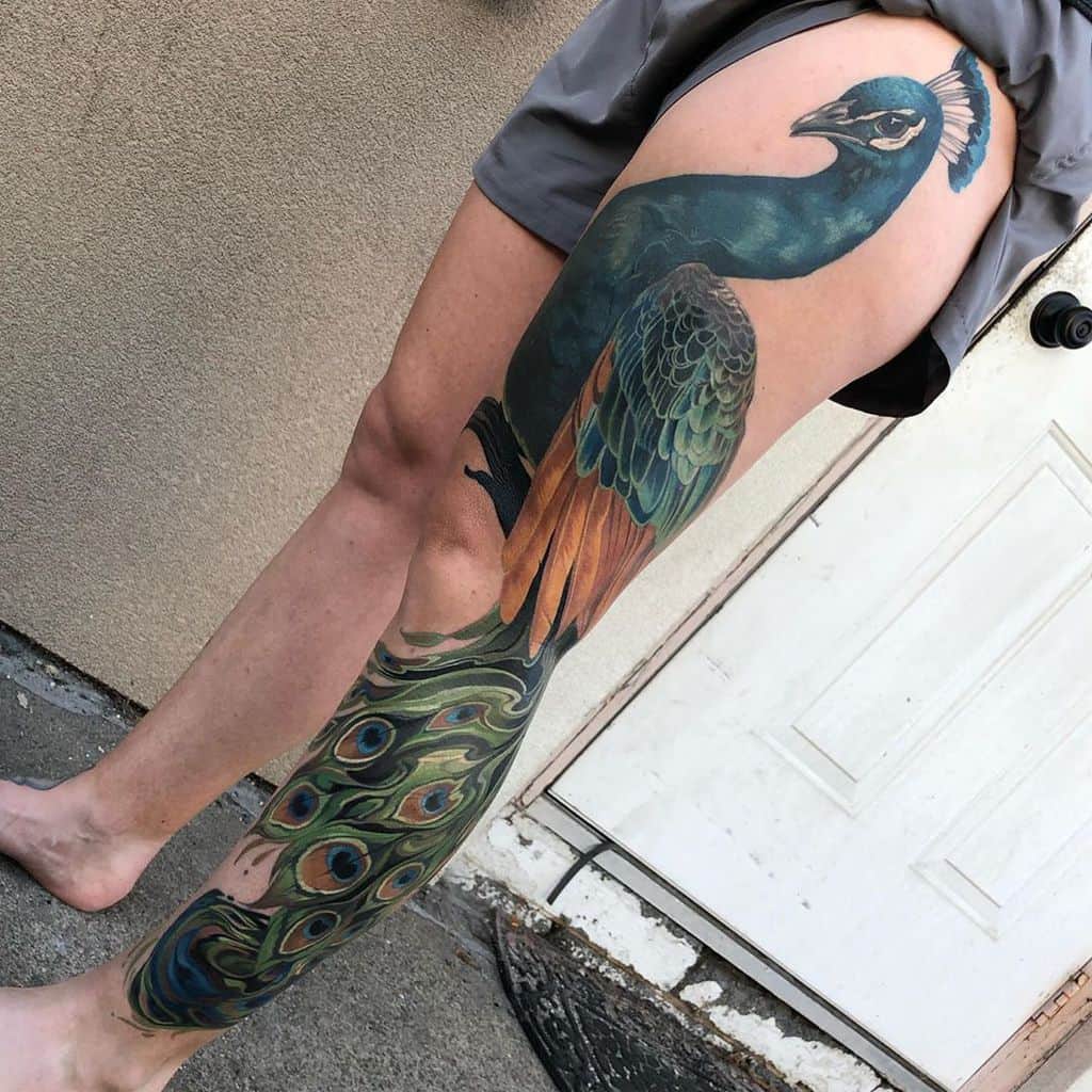 Full-length peacock tattoo on the side of the leg with vibrant blue and green feathers, showing the peacock's intricate tail feathers and colorful details.