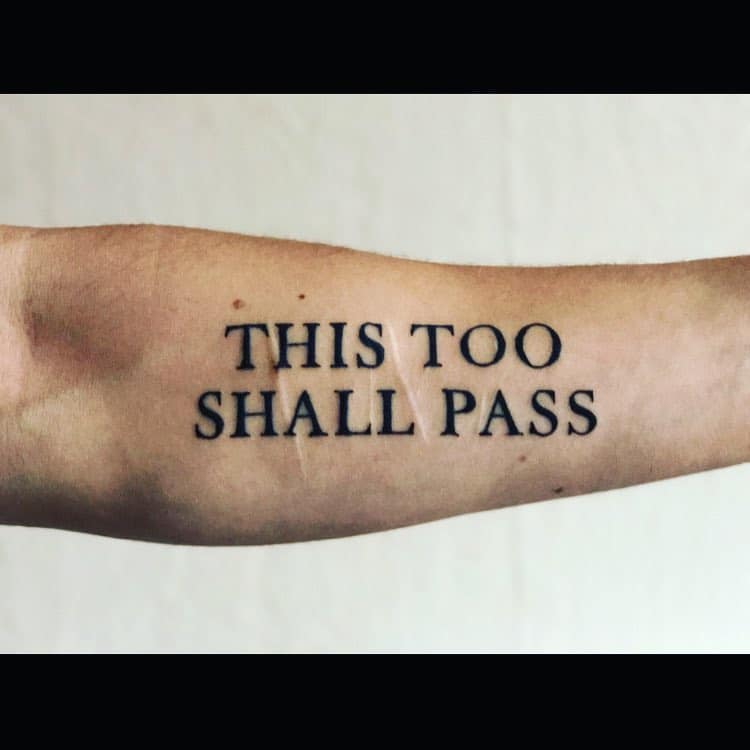 A "This Too Shall Pass" tattoo on a person's forearm, with bold, uppercase letters.