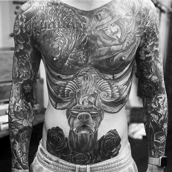 Full torso black and grey tattoo featuring a central Doberman with a muzzle, surrounded by roses, an eagle, and detailed mythological and animal imagery across the chest and arms.