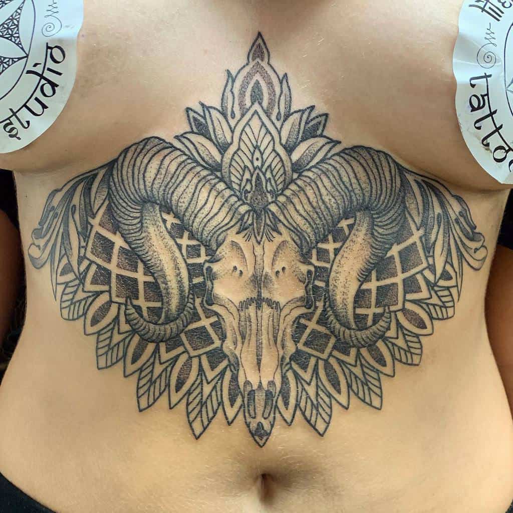 A detailed tattoo of a ram skull with intricate patterns on a woman's stomach showcases deep artistry