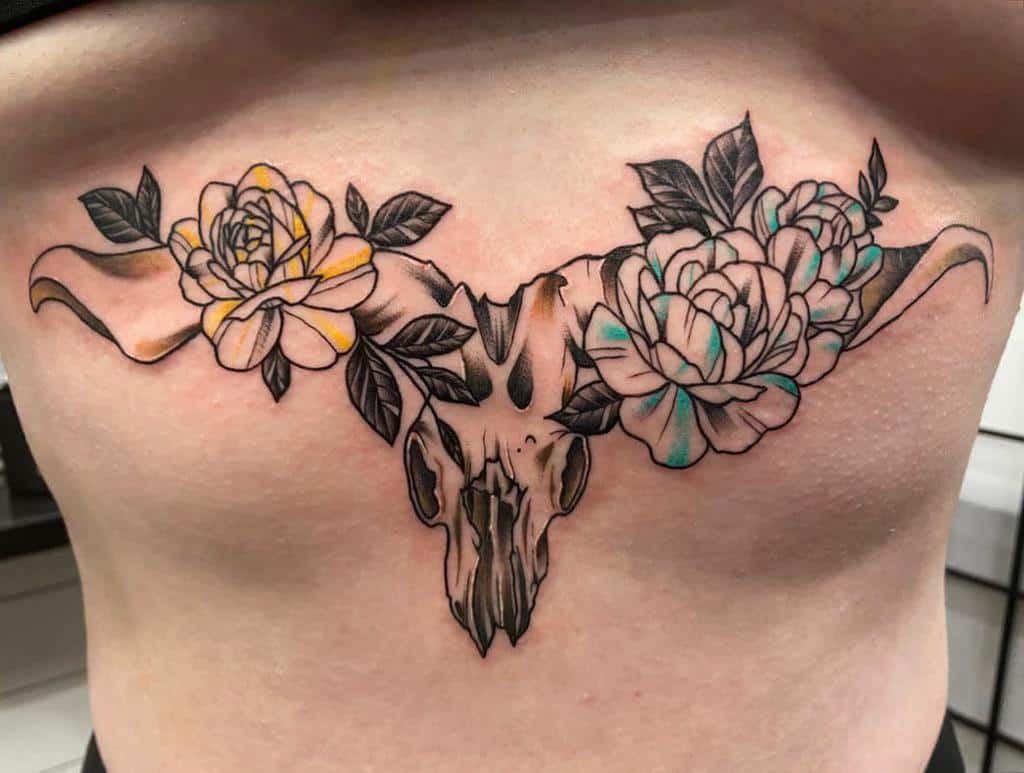 Tattoo of a ram skull adorned with two flowers and leaves on either side, located on the stomach