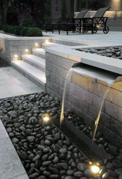 Modern backyard waterfall with sleek design, black pebbles, and integrated lighting.