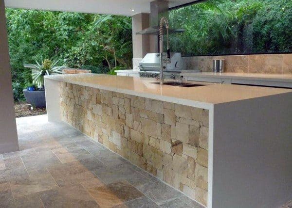 Modern outdoor kitchen with stone island, sleek countertop, and grill, surrounded by lush outdoor greenery