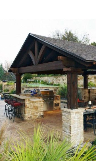 Outdoor BBQ bar with a stone foundation, rich wooden beams, and a gable roof in a spacious patio area.