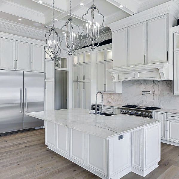 Elegant white kitchen with marble backsplash, matching island, intricate pendant lighting, and stainless steel appliances