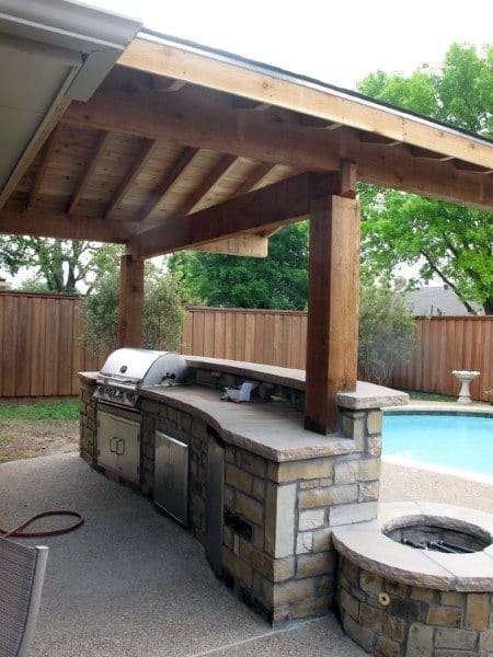 Outdoor kitchen with a grill under a wooden pergola, next to an outdoor pool and wooden fence
