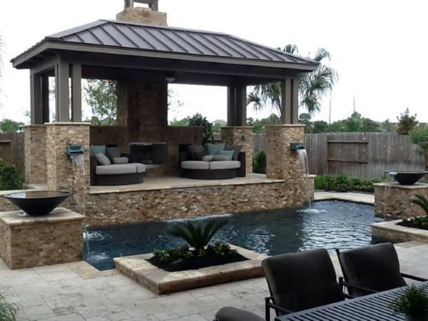 Elevated stone pavilion with a metal roof, cozy seating, and water features surrounding a tranquil pool.