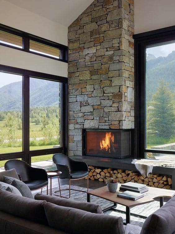 Living room with stone corner fireplace, large windows, modern seating, and stacked firewood storage.