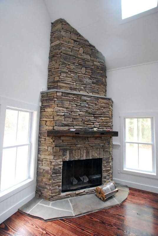 Rustic stone corner fireplace with wooden mantel, tall chimney, and hardwood flooring.