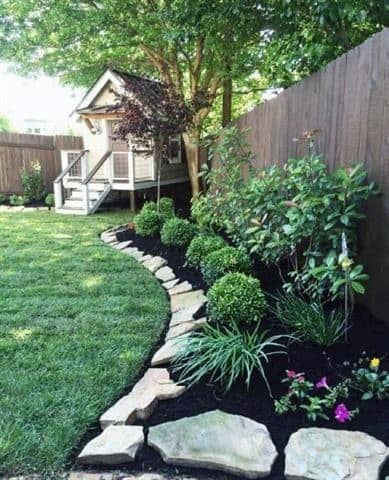 A curved stone border along a garden bed with lush greenery and colorful flowers, leading to a small garden shed