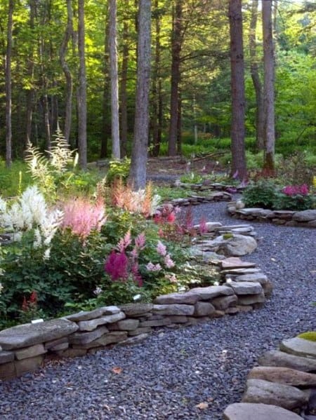 A stone-bordered garden path winding through a forest, with colorful flowers and gravel filling the beds along the way