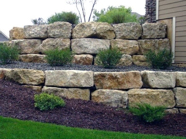 A large stone retaining wall made of stacked boulders, creating multiple terraced levels with shrubs and mulch
