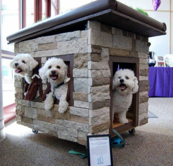 Dog house with a stone exterior and multiple windows, featuring three dogs peeking out, set on a rolling base with a roof