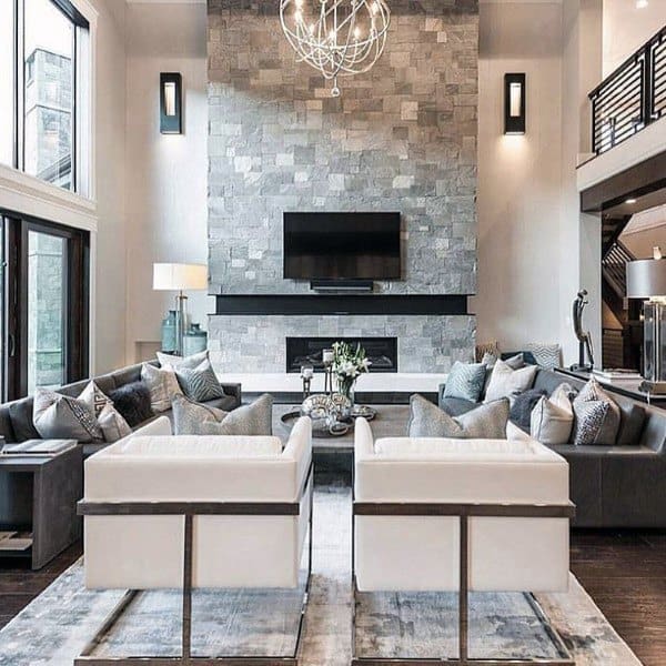 Modern living room with a tall stone fireplace, chandelier, and sleek gray and white furniture.