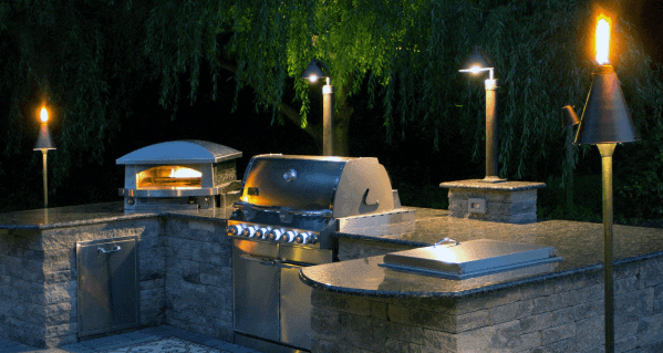 Built-in grill and pizza oven in an outdoor kitchen illuminated by lights, nestled in lush greenery at night