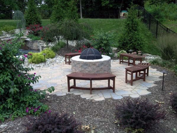 A stone fire pit with wooden benches surrounded by greenery in a landscaped garden