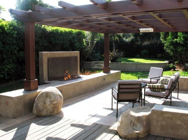 Cozy patio with chairs and a fire pit under a pergola, enveloped in greenery and large stone accents
