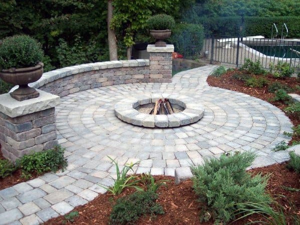 Circular stone patio with fire pit, surrounded by greenery and a curved stone bench, pool in the background