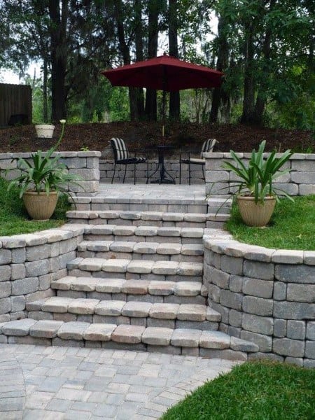 Stone retaining wall with steps leading to a patio, featuring potted plants and a red umbrella.