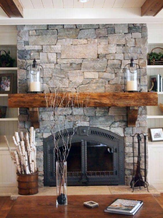 Stone fireplace with wooden mantel, lantern candles, birch logs, and rustic decor elements.