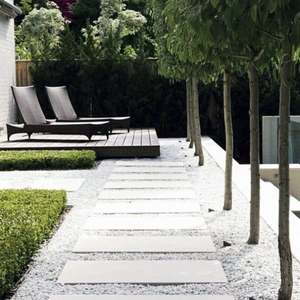 Modern backyard with stone slab walkway, gravel, manicured hedges, and lounge chairs on a deck.