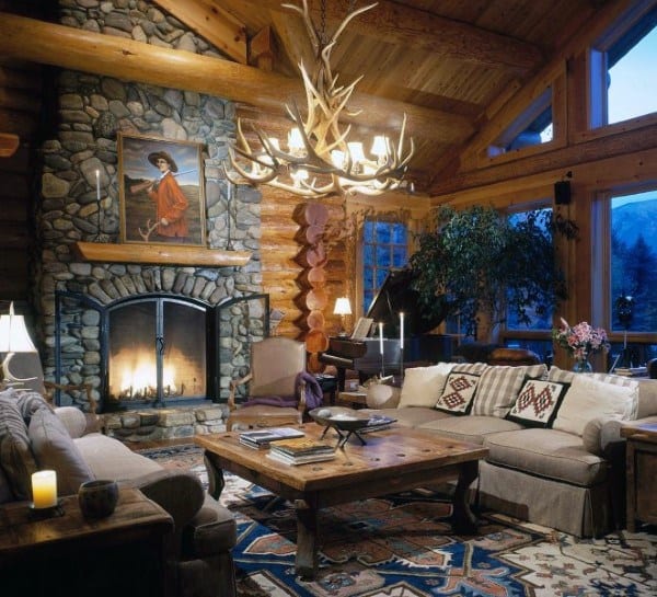 Rustic log cabin living room with a stone fireplace, antler chandelier, and cozy wooden accents.