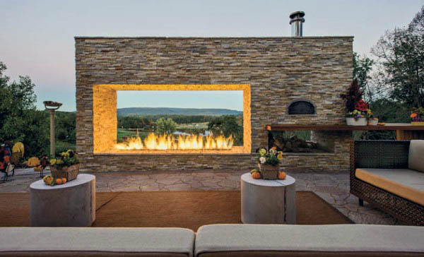 Outdoor patio with a large stone fireplace, surrounded by seating and trees, overlooking a scenic view