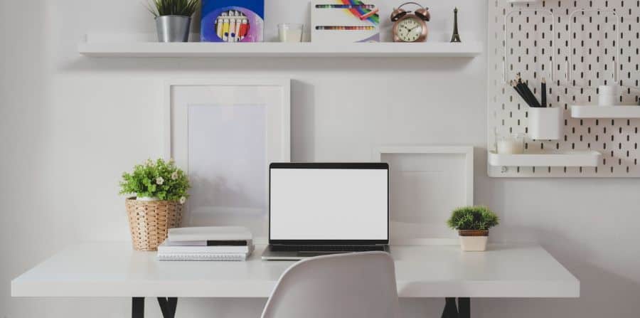 Simple white desk setup with laptop, plants, and wall shelves for organization.