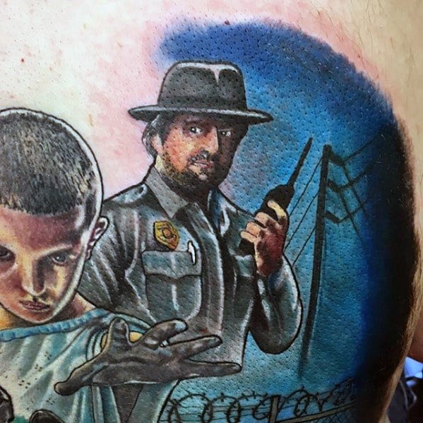 Colorful Stranger Things tattoo of Eleven using her powers beside Chief Hopper holding a radio, with a dramatic sky and barbed wire in the background.