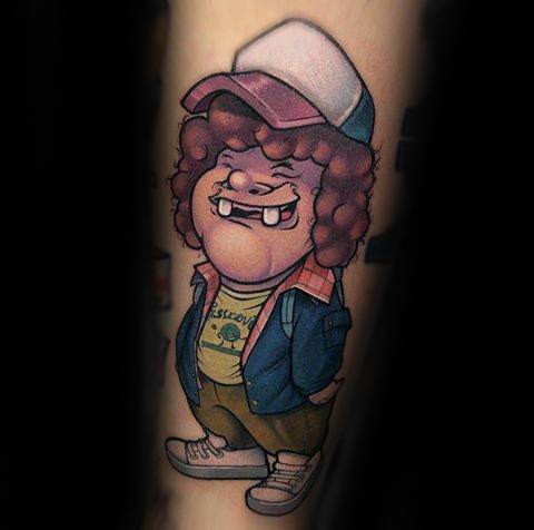 Cartoon-style tattoo of Dustin from Stranger Things, smiling confidently in his signature outfit with cap, curly hair, and graphic tee.