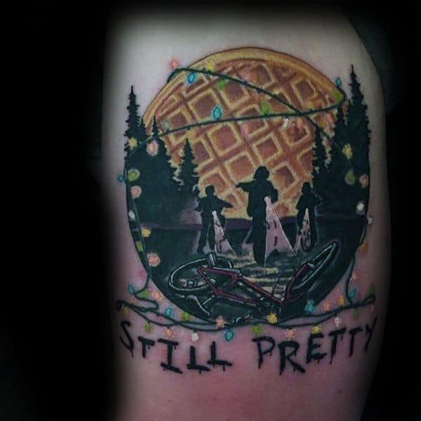 Stranger Things tattoo featuring kids walking in the woods under Christmas lights, a giant waffle moon, and the phrase “Still Pretty” below.
